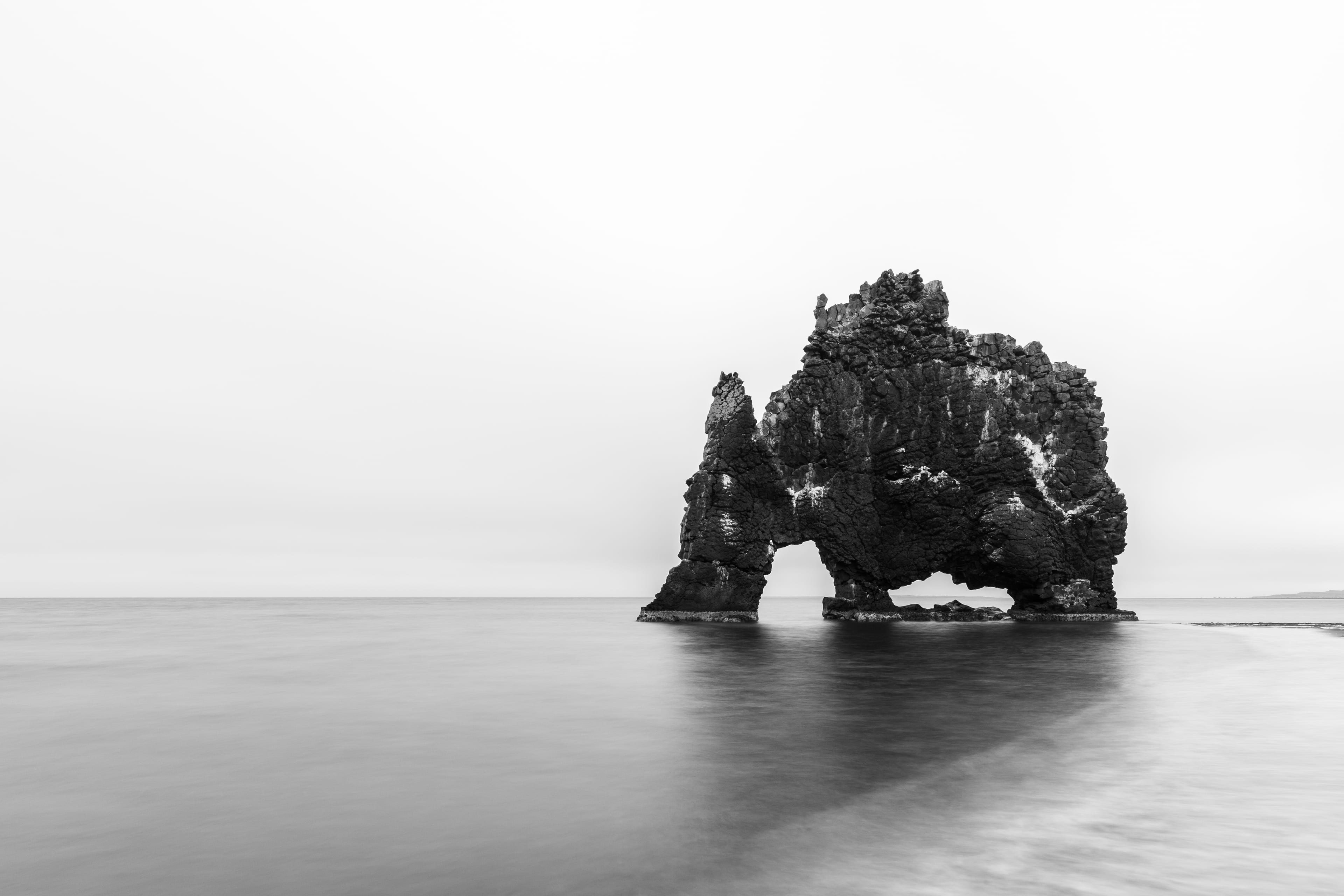 Un rocher de basalte isolé dans une mer étale, photographié en noir et blanc en pose longue sous un ciel presque blanc